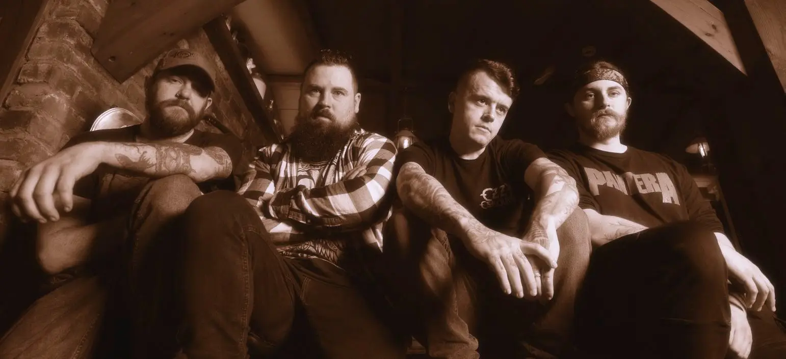 Four men sit closely together in a dim, sepia-toned attic setting, looking at the camera with serious expressions (likely a band portrait).