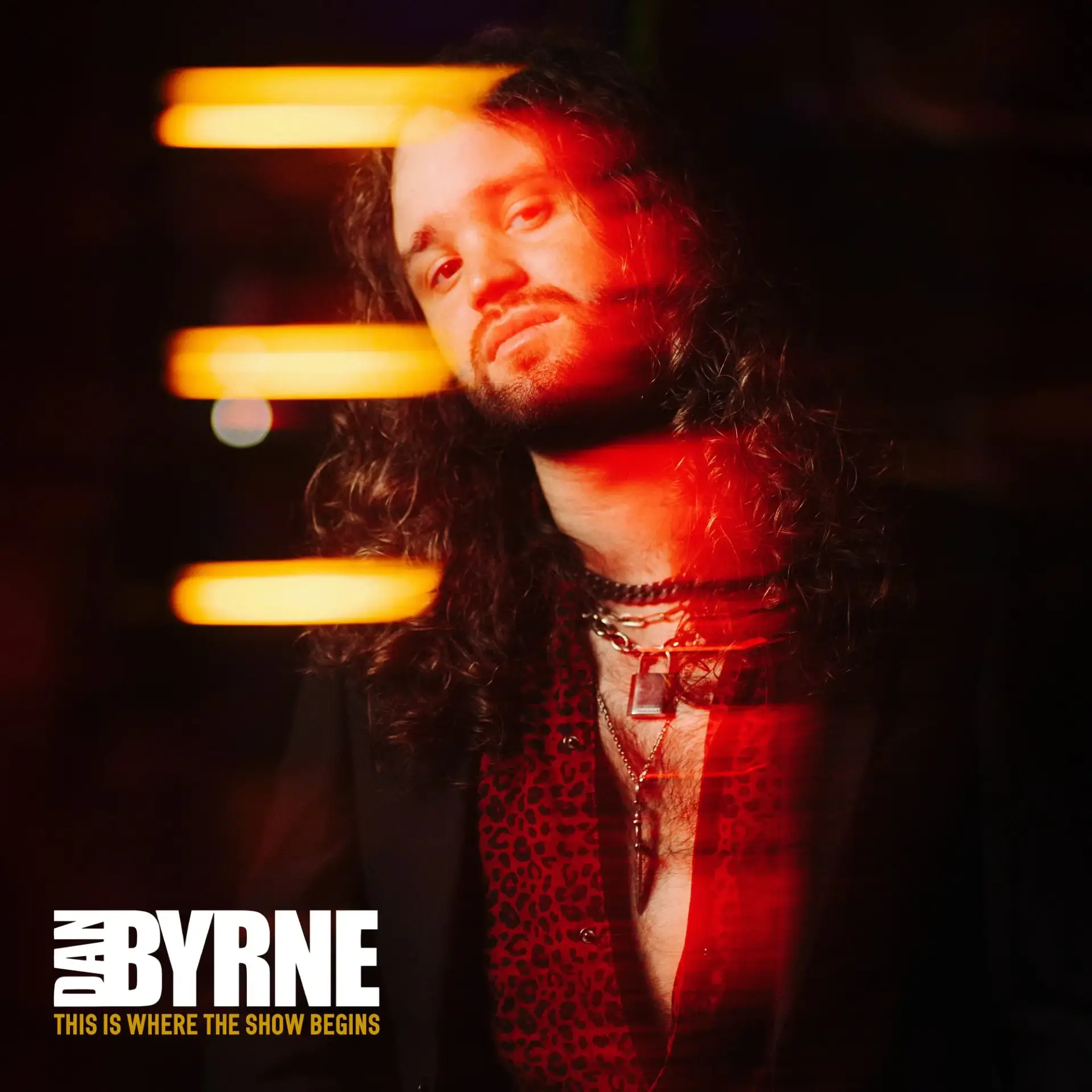 Close-up portrait of a man with long hair under red stage lighting; large white text reads 'BYRNE' with a yellow subtitle below.