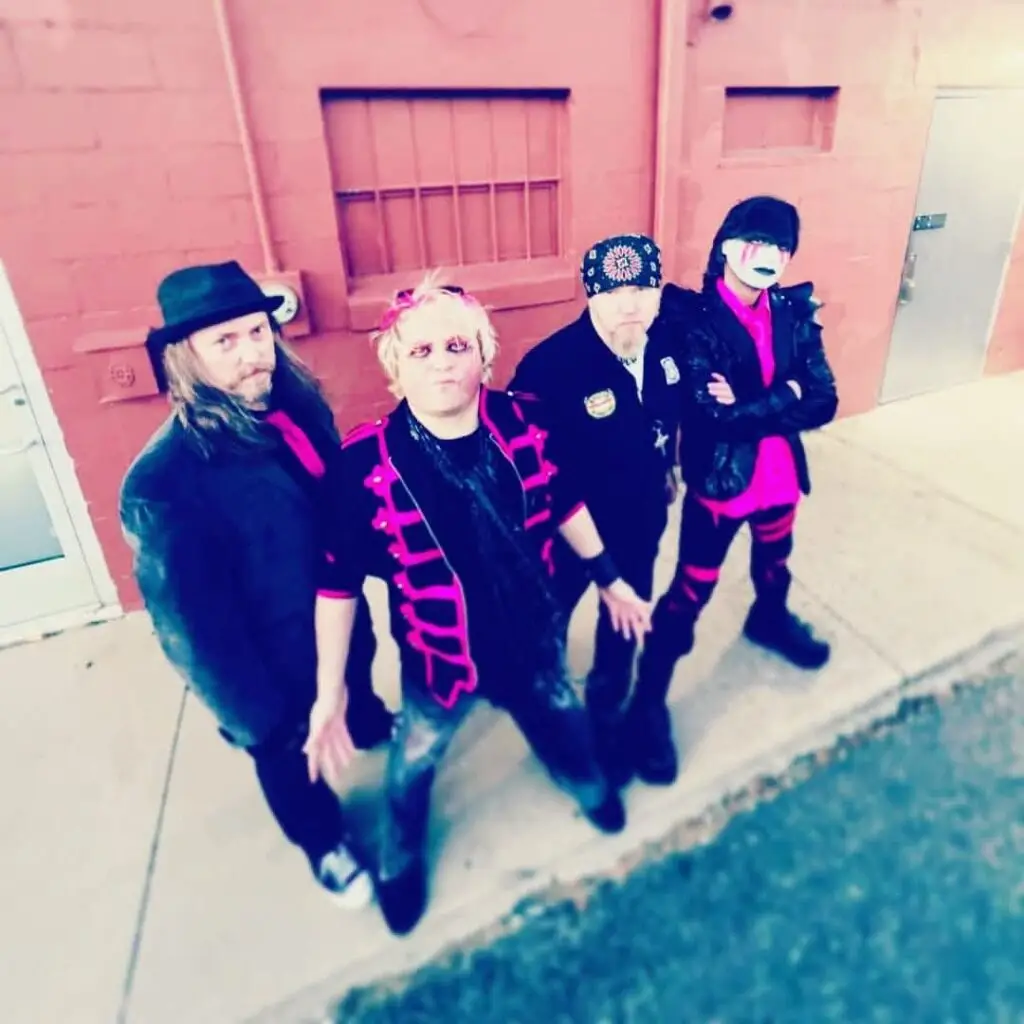 Four musicians in bold pink-and-black outfits stand on a sidewalk against a red brick wall, posing for a photo; one wears a black hat, another with bright pink hair, and a fourth with a white-painted face mask.