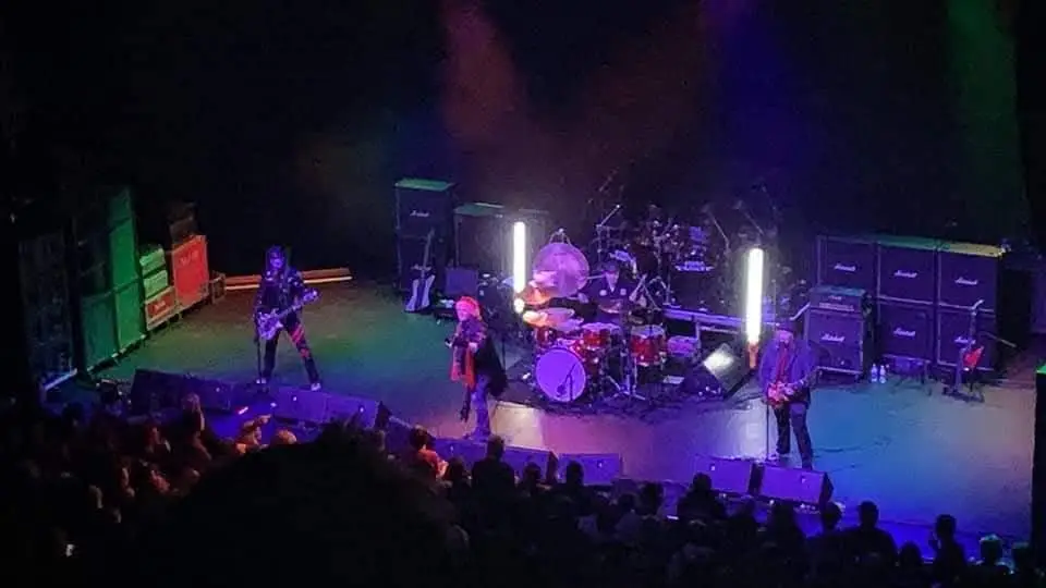 A five‑piece rock band performs on a dimly lit stage with amplifiers, drums, and colorful lights, while a crowd watches from the foreground.