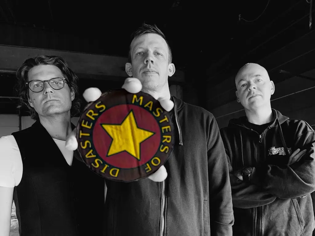 Three men pose indoors; the central man extends a round badge reading 'Masters of Disaster' with a yellow star toward the camera.