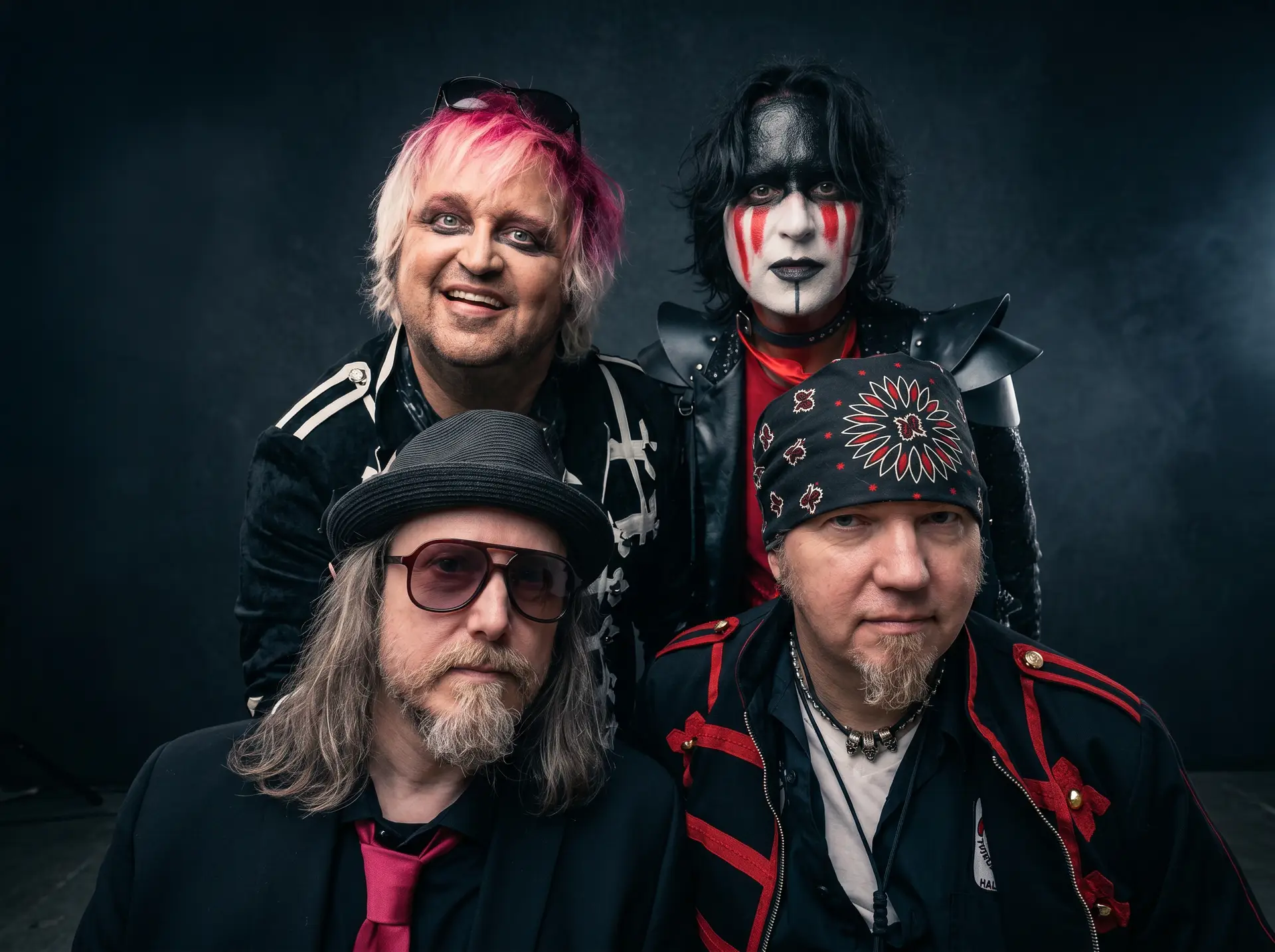 Group portrait of four men in theatrical punk/goth costumes with face paint and accessories, posing against a dark studio backdrop.