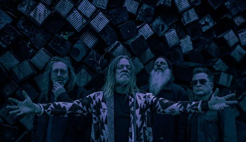 Four men pose in front of a dark, geometric metal wall; center figure extends arms wide, wearing a patterned jacket.