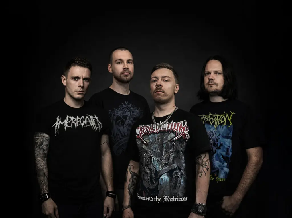 Four male musicians in black band shirts pose together against a dark background, a metal/rock group portrait.