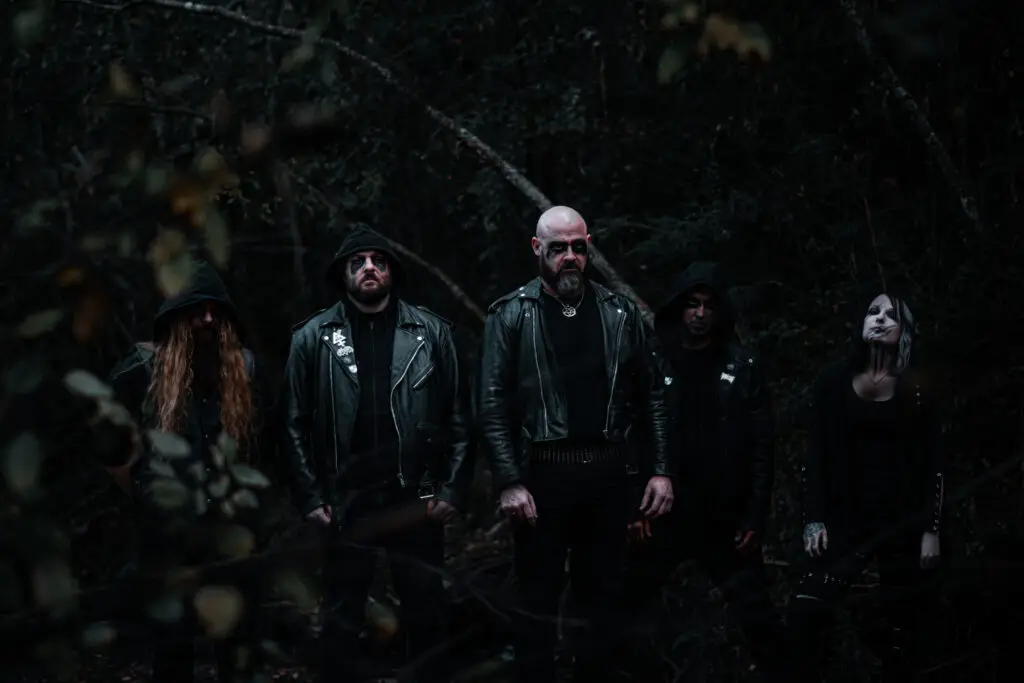 Five people in black leather jackets with dark makeup stand in a dim, wooded area, looking at the camera.