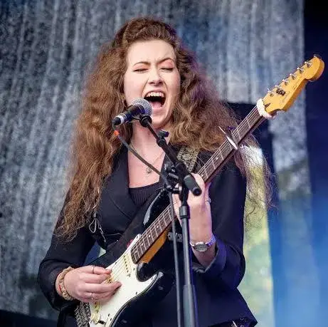 Female guitarist and vocalist passionately singing into a microphone while playing an electric guitar on stage with a dark blazer and curly hair lighted by stage lighting.