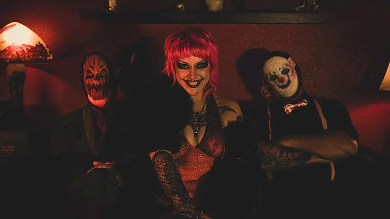 Nova Riot - the Riff Report Blog Three adults in horror masks sit on a dark red couch; center figure has bright red hair and a smiling, theatrical expression.