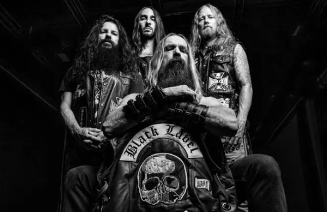 Group portrait of four long-haired metal musicians in leather, front man crossing arms with a Black Label patch featuring a skull logo on his vest.
