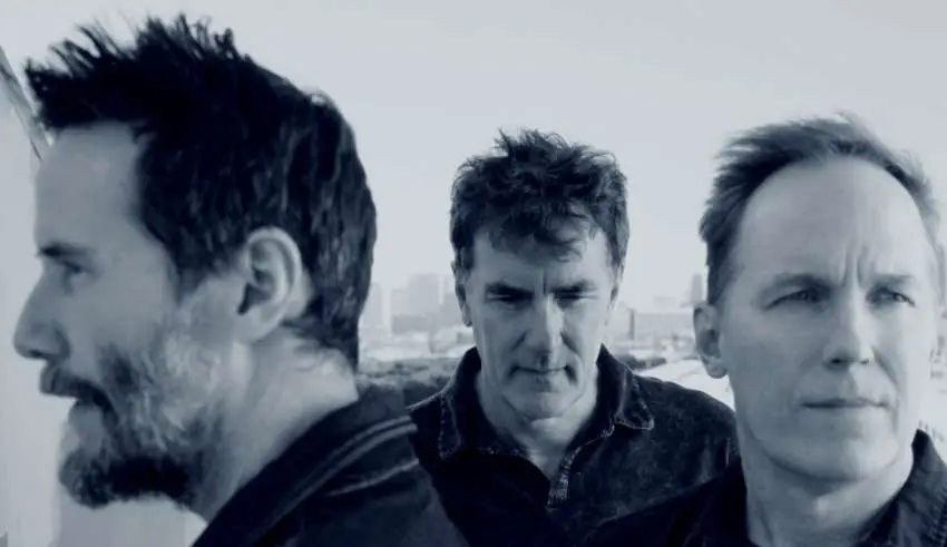 Three men stand close together outdoors, with a distant city skyline in the background.