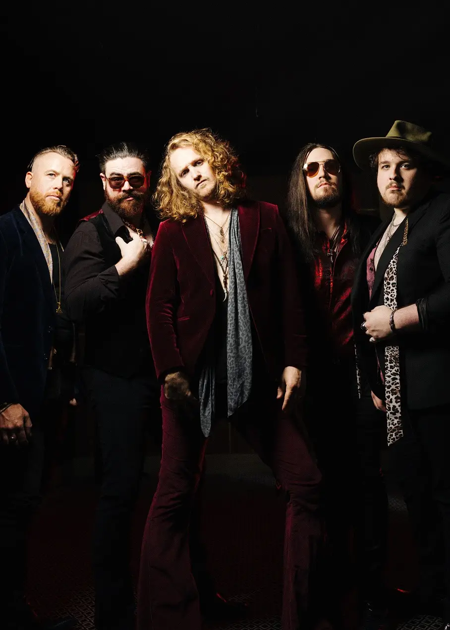 Five male musicians posing together in a dimly lit studio, forming a rock band portrait.