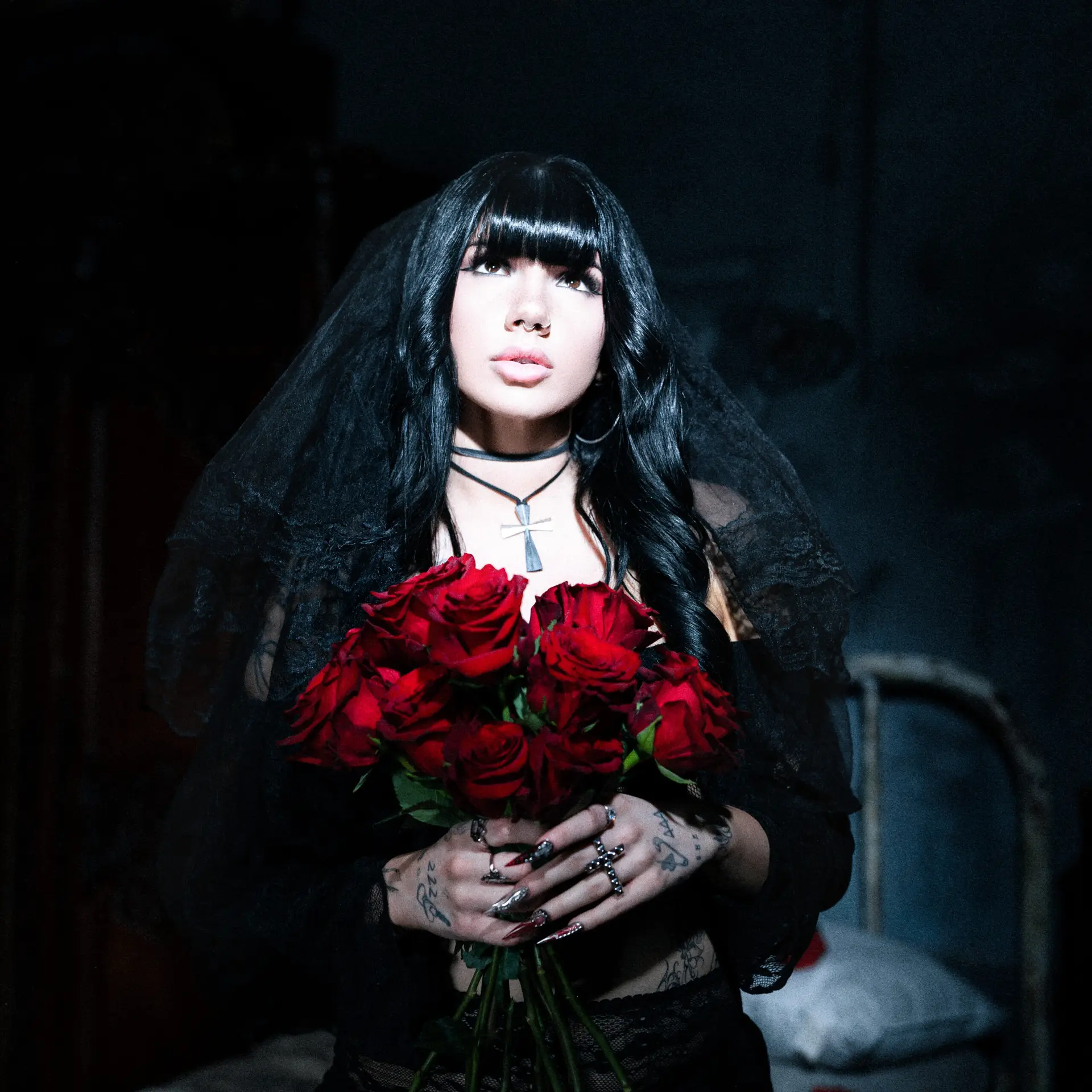 Portrait of a woman with long black hair and a lace veil, holding a bouquet of red roses against a dark background.