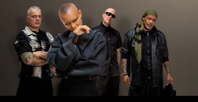 Group of four men in dark outfits posing together against a neutral backdrop, looking confident.