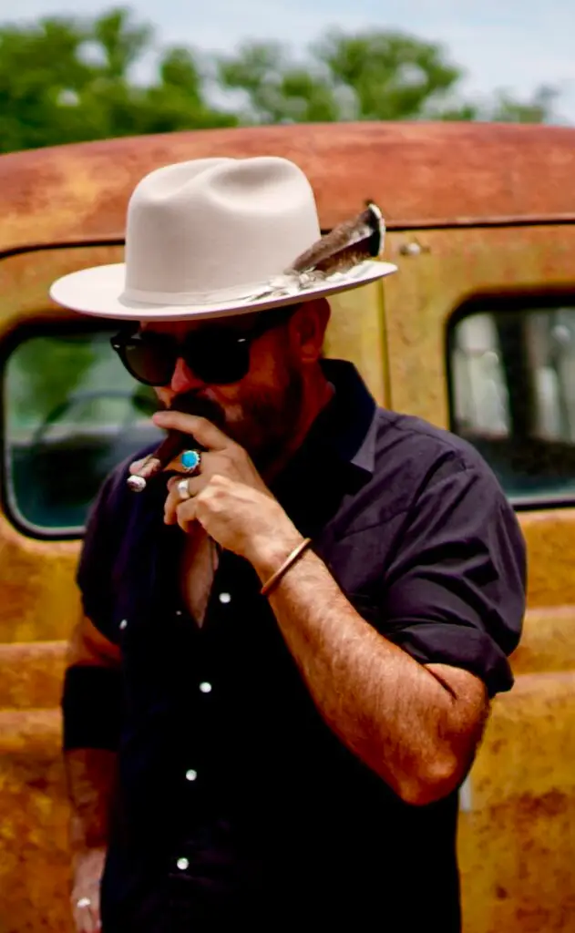 Steve Harris Cigar - Photo Credit = Joe Ward - the Riff Report Blog Man wearing a white fedora and sunglasses, smoking a cigar beside a rusted truck.