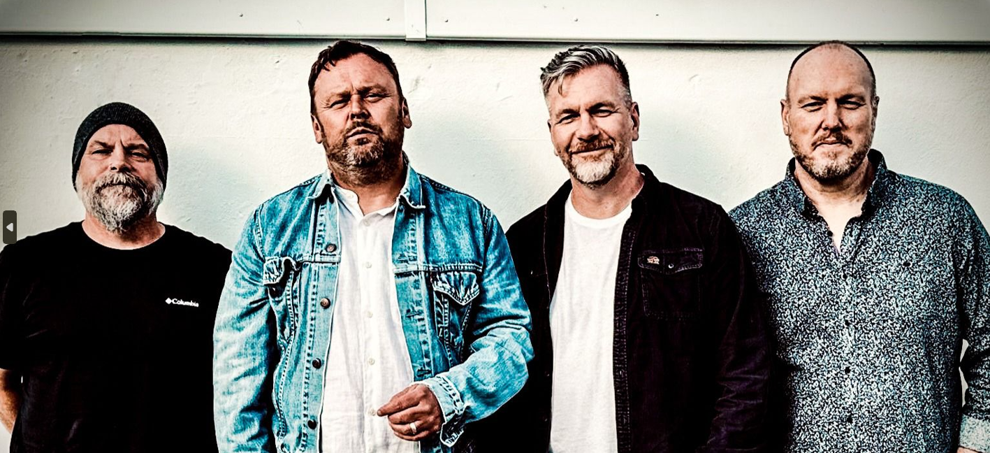 Four men stand in a row against a light wall, wearing casual outfits (denim jacket, button-down, and patterned shirt).