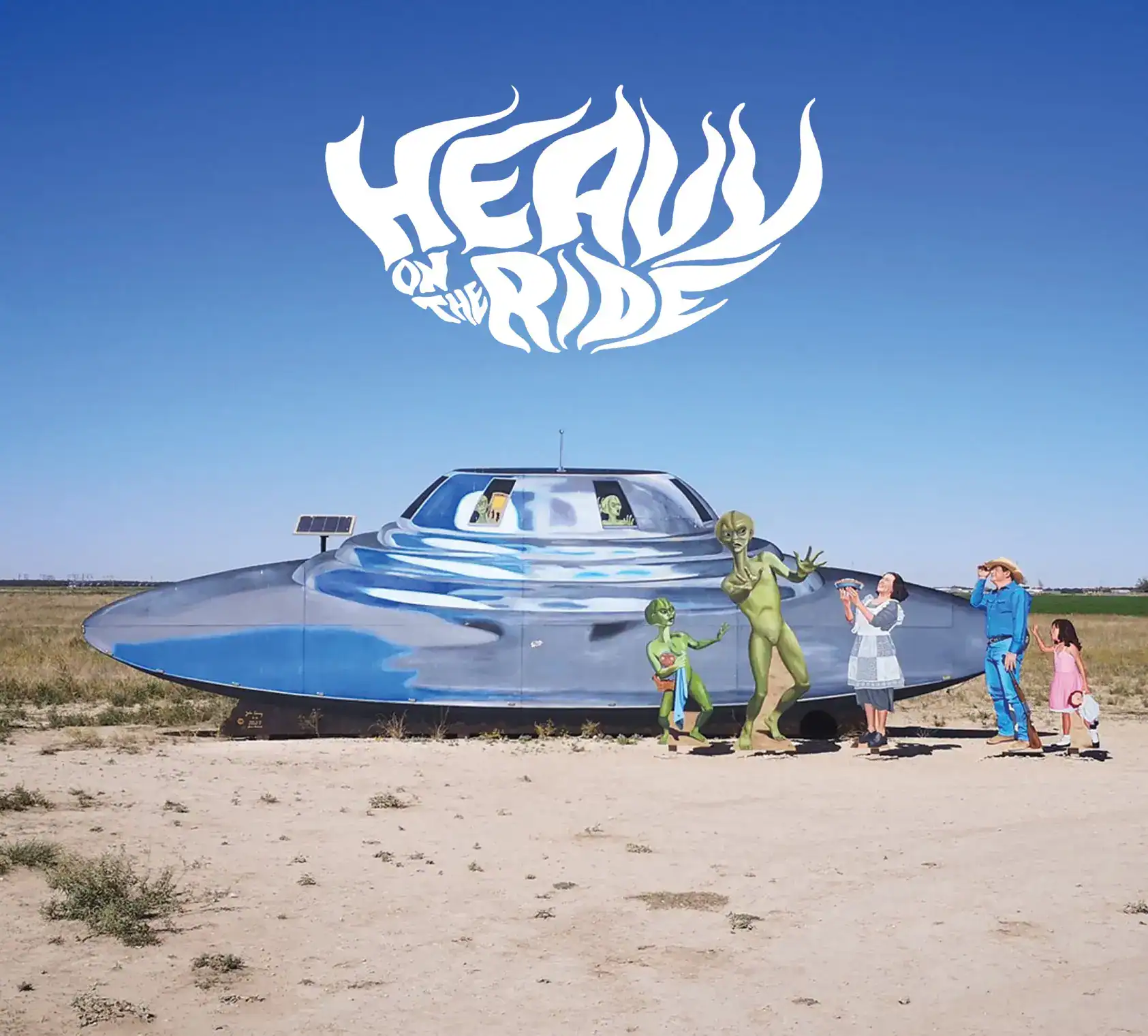 Desert scene with a large blue-painted flying-saucer mural and life-size cartoon aliens posing with a family and visitors nearby.