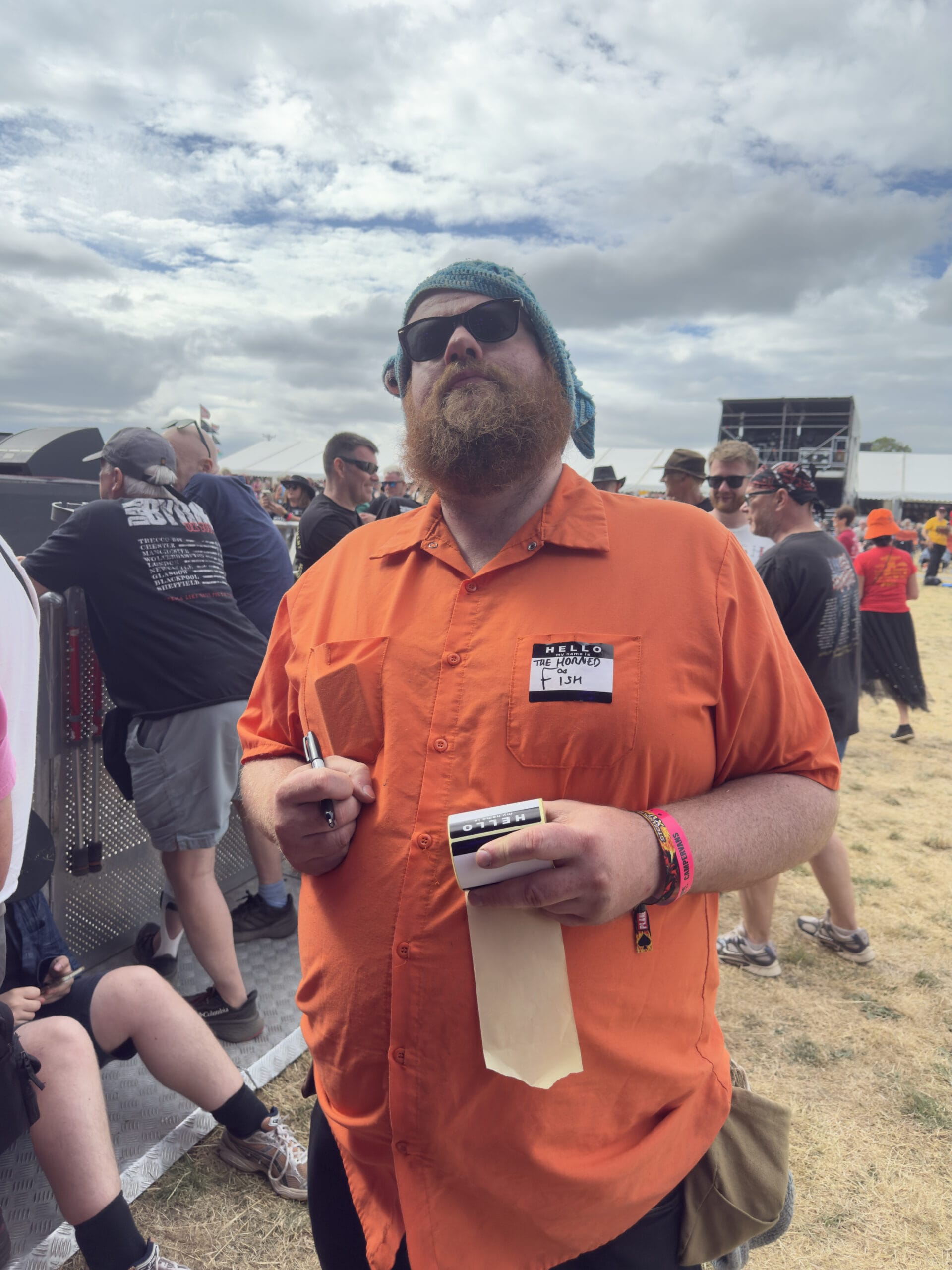 Bearded man in an orange shirt with a teal headscarf and sunglasses at an outdoor festival, holding a roll of paper and a marker.
