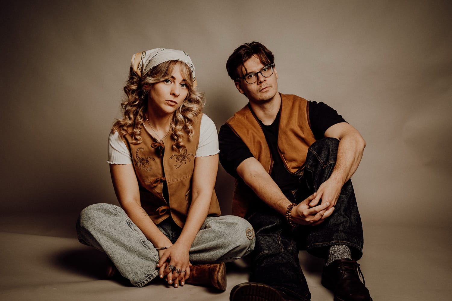 Two young adults in vintage western outfits sit crossโlegged on the floor, looking at the camera against a neutral backdrop.